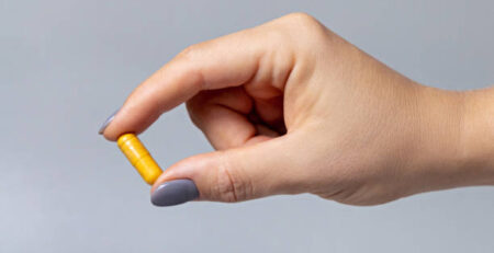 Mão feminina segurando uma cápsula amarela com os dedos em close-up sobre fundo cinza. Conceitos de saúde, médicos e farmacêuticos.